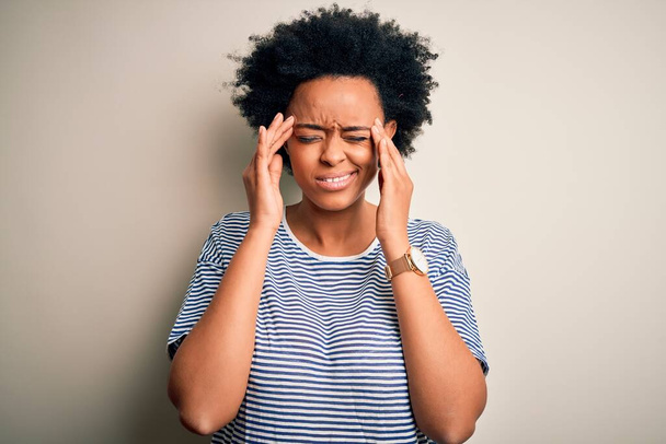 Junge schöne afroamerikanische Afro-Frau mit lockigem Haar trägt gestreiftes T-Shirt mit der Hand auf dem Kopf für Kopfschmerzen wegen Stress. Migräne. - Foto, Bild