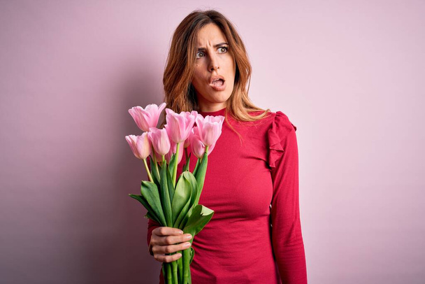 Mladá krásná brunetka žena drží kytici růžových tulipánů na izolovaném pozadí v šoku tvář, vypadá skeptický a sarkastický, překvapený s otevřenými ústy - Fotografie, Obrázek