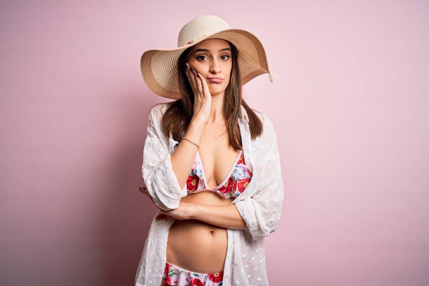 Giovane bella donna in vacanza indossa bikini e cappello estivo su sfondo rosa pensando cercando stanco e annoiato con problemi di depressione con le braccia incrociate
. - Foto, immagini