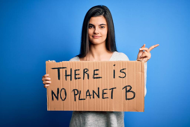 jong mooi brunette activist vrouw houden spandoek protesteren naar zorg de planeet zeer gelukkig wijzend met hand en vinger naar de zijkant - Foto, afbeelding