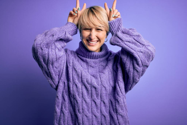 Nuori blondi nainen lyhyet hiukset yllään talvi poolopaita villapaita violetti tausta Poseeraa hauska ja hullu sormet pään kuin pupu korvat, hymyillen iloinen
 - Valokuva, kuva