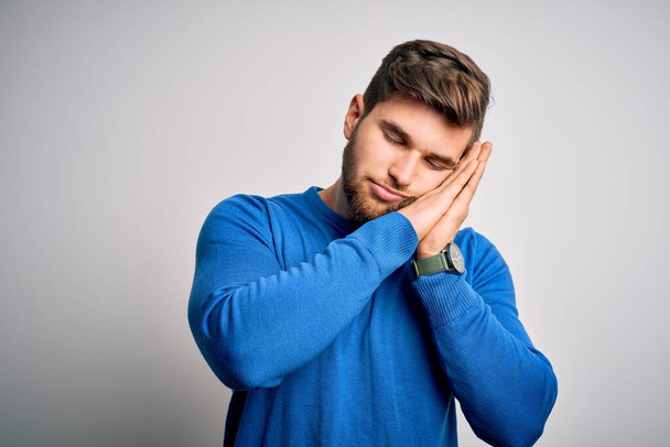 Young handsome blond man with beard and blue eyes wearing casual sweater sleeping tired dreaming and posing with hands together while smiling with closed eyes. - Foto, Imagen