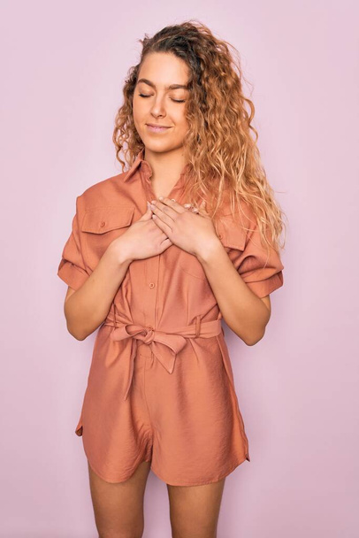 Young beautiful woman with blue eyes wearing casual summer dress over pink background smiling with hands on chest with closed eyes and grateful gesture on face. Health concept. - 写真・画像