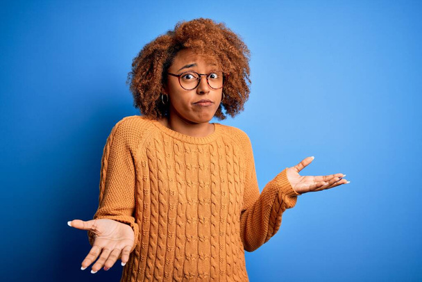 Junge schöne afroamerikanische Afro-Frau mit lockigem Haar trägt lässigen Pullover ahnungslos und verwirrt Ausdruck mit erhobenen Armen und Händen. Zweifelhaftes Konzept. - Foto, Bild