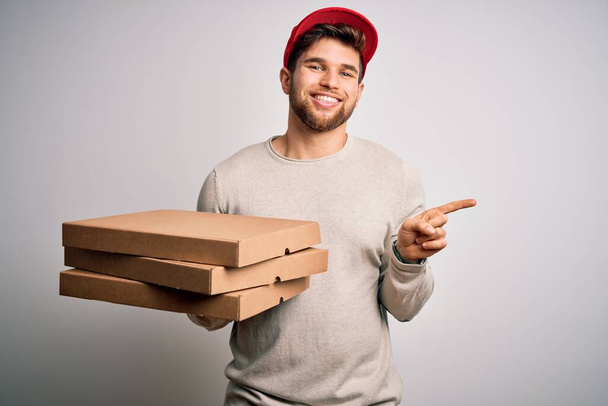 Jonge blonde bezorger met baard en blauwe ogen met kartonnen pizza heel gelukkig wijzend met hand en vinger naar de zijkant - Foto, afbeelding