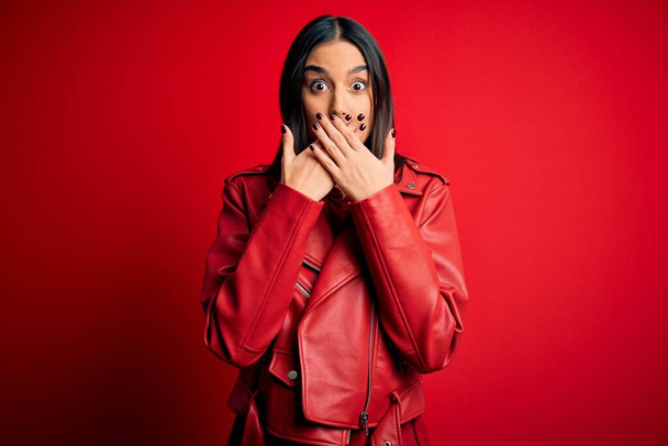 Young beautiful brunette woman wearing casual jacket standing over isolated red background shocked covering mouth with hands for mistake. Secret concept. - Zdjęcie, obraz