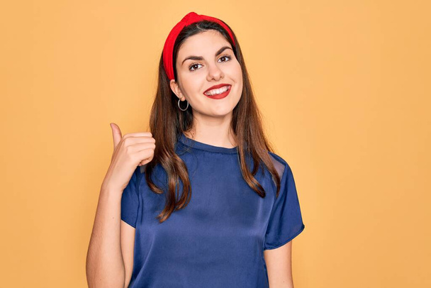 Young beautiful brunette woman wearing red lips over yellow background smiling with happy face looking and pointing to the side with thumb up. - Photo, Image
