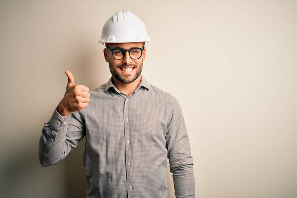 Молодий архітектор чоловік в шоломі будівельника на ізольованому фоні робить щасливі великі пальці вгору жестом рукою. Затвердження виразу, який показує успіх фотоапарата
. - Фото, зображення