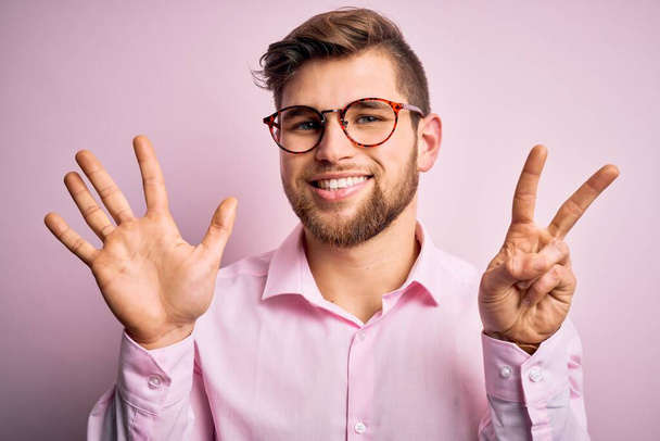 Mladý pohledný blonďák s vousy a modrýma očima v růžové košili a brýlích ukazující a ukazující prsty číslo sedm a usmívající se sebevědomě a šťastně. - Fotografie, Obrázek