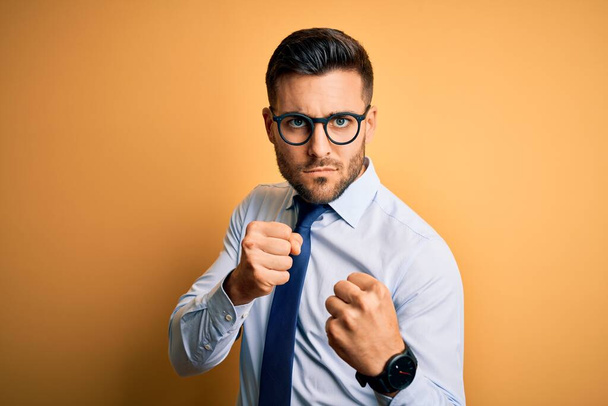 Young handsome businessman wearing tie and glasses standing over yellow background Ready to fight with fist defense gesture, angry and upset face, afraid of problem - Photo, Image
