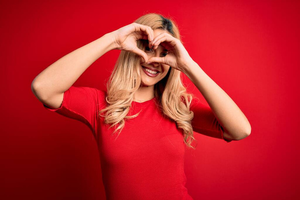 Молодая красивая блондинка в повседневной футболке стоит на изолированном красном фоне и делает форму сердца с рукой и пальцами, улыбаясь, глядя сквозь знак
 - Фото, изображение