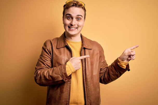 Junger hübscher rothaariger Mann in lässiger Lederjacke vor isoliertem gelben Hintergrund, lächelnd und mit zwei Händen und Fingern zur Seite in die Kamera blickend. - Foto, Bild