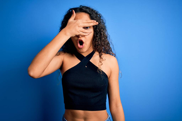 Young beautiful woman with curly hair wearing casual t-shirt standing over blue background peeking in shock covering face and eyes with hand, looking through fingers with embarrassed expression. - Fotoğraf, Görsel