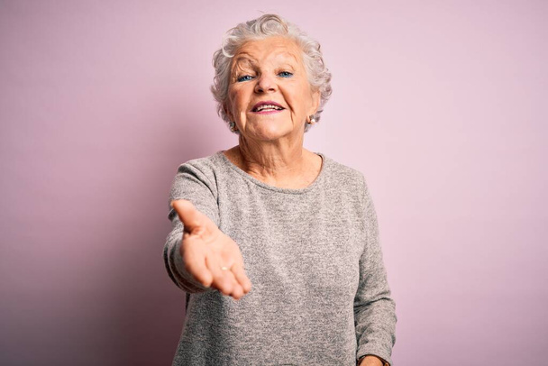 Старшая красивая женщина в повседневной футболке стоя на изолированном розовом фоне улыбаясь весело предлагая ладонь помощи и принятия
. - Фото, изображение