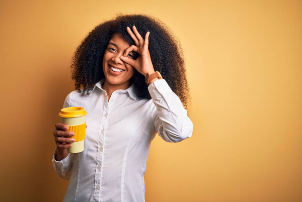 Młoda afrykańska kobieta biznesu z afro włosy picie kawy z zabrać filiżankę z uśmiechem szczęśliwej twarzy robi ok znak z ręki na oko patrząc przez palce - Zdjęcie, obraz