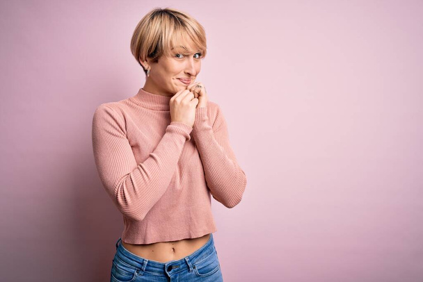 Mujer rubia joven con el pelo corto con suéter casual cuello alto sobre fondo rosa riendo nerviosa y emocionada con las manos en la barbilla mirando a un lado
 - Foto, imagen