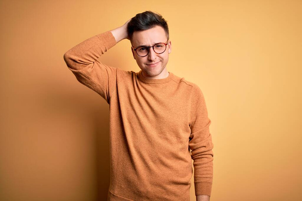 Ein junger gutaussehender kaukasischer Mann mit Brille und lässigem Winterpullover vor gelbem Hintergrund verwirrt und fragt sich, was das soll. Unsicher im Zweifel, mit der Hand auf dem Kopf denkend. Umfassendes Konzept. - Foto, Bild
