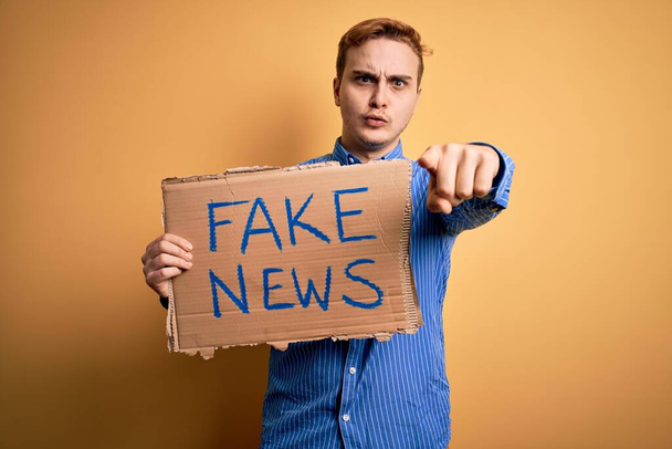 Junger hübscher rothaariger Mann mit Transparent mit Fake-News-Botschaft über gelbem Hintergrund, der mit dem Finger in die Kamera und zu dir zeigt, selbstbewusste Geste, die ernst aussieht - Foto, Bild