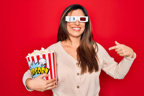 Young hispanic woman wearing 3d movie glasses and eating popcorn over red background with surprise face pointing finger to himself - Foto, Imagen
