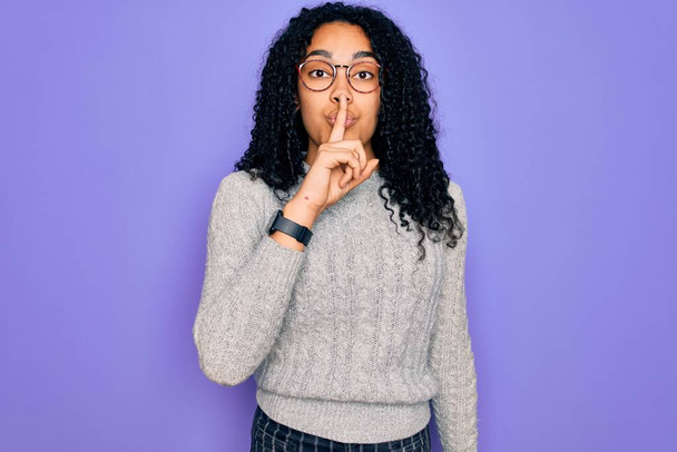 Mujer afroamericana joven con suéter casual y gafas sobre fondo púrpura pidiendo estar en silencio con el dedo en los labios. Silencio y concepto secreto
. - Foto, imagen