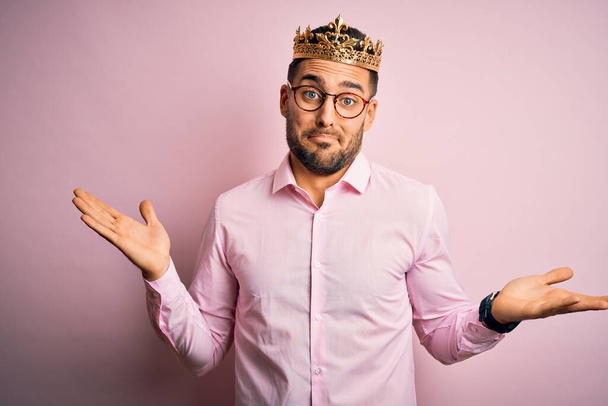 Jonge knappe zakenman draagt gouden kroon als koning over roze achtergrond clueless en verwarde uitdrukking met armen en handen opgeheven. Twijfelachtig concept. - Foto, afbeelding