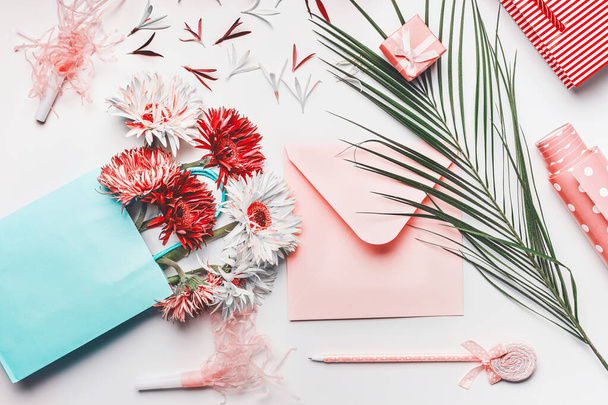 Female greeting objects flat lay in coral and turquoise colors. Shopping bag with Flowers bunch, blank envelope with pen, palm leaves and wrapping paper on white background, top view. - Photo, Image