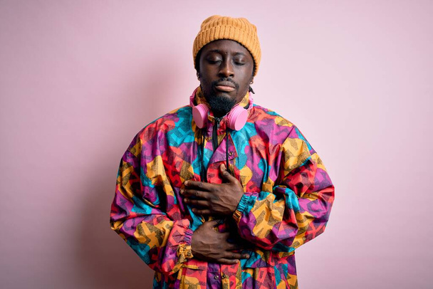 Νεαρός όμορφος Αφροαμερικάνος που φοράει πολύχρωμο παλτό και καπέλο πάνω από ροζ φόντο με το χέρι στο στομάχι επειδή δυσπεψία, επώδυνη ασθένεια αισθάνεται άσχημα. Έννοια Ache. - Φωτογραφία, εικόνα