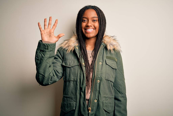 Joven mujer afroamericana vistiendo abrigo de parka de invierno sobre fondo aislado mostrando y señalando hacia arriba con los dedos número cinco mientras sonríe confiado y feliz
. - Foto, Imagen