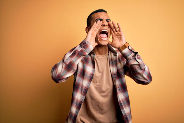 Young handsome african american man wearing casual shirt standing over yellow background Shouting angry out loud with hands over mouth - Foto, imagen