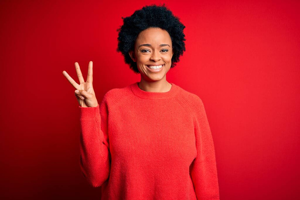 Νεαρή όμορφη Αφρο-Αμερικανίδα γυναίκα με σγουρά μαλλιά φορώντας casual πουλόβερ που δείχνει και δείχνει προς τα πάνω με τα δάχτυλα νούμερο τρία, ενώ χαμογελά αυτοπεποίθηση και χαρούμενος. - Φωτογραφία, εικόνα