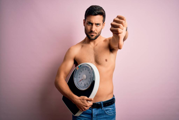 Jonge knappe slanke man met baard shirtloze houden gewicht machine over roze achtergrond met boos gezicht, negatief teken toont een hekel aan met duimen naar beneden, afwijzing concept - Foto, afbeelding