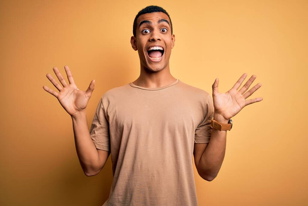 Young handsome african american man wearing casual t-shirt standing over yellow background celebrating crazy and amazed for success with arms raised and open eyes screaming excited. Winner concept - Valokuva, kuva
