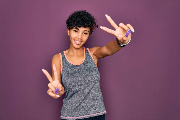 Junge schöne afrikanisch-amerikanische Afro-Sportlerin beim Turnen in Sportbekleidung, lächelnd mit herausgestreckter Zunge, die Finger beider Hände beim Siegeszeichen zeigt. Nummer zwei. - Foto, Bild
