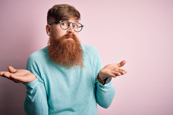 Bonito irlandês ruiva homem com barba vestindo óculos sobre rosa isolado fundo indolente e confuso expressão com braços e mãos levantadas. Conceito de dúvida
. - Foto, Imagem
