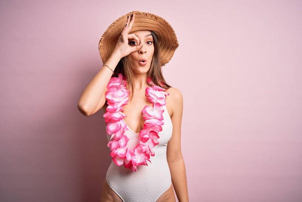 Mladá krásná bruneta žena na dovolené v plavkách a havajských květinách lei dělat ok gesto šokován s překvapeným obličejem, oko dívá skrz prsty. Nevěřící výraz. - Fotografie, Obrázek