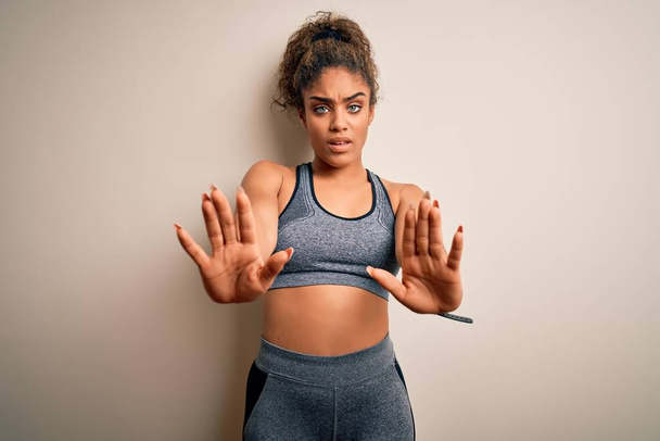 Молодая африканская американская спортсменка, занимающаяся спортом в спортивной одежде на белом фоне делает остановку жест с ладонями рук, сердитый и разочарование выражение
 - Фото, изображение