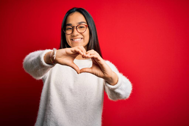 Młoda piękna azjatycka kobieta ubrana w luźny sweter i okulary na czerwonym tle uśmiechnięta w miłości robi kształt symbolu serca rękami. Romantyczna koncepcja. - Zdjęcie, obraz