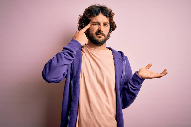 Young handsome sporty man with beard wearing casual sweatshirt over pink background confused and annoyed with open palm showing copy space and pointing finger to forehead. Think about it. - Photo, Image