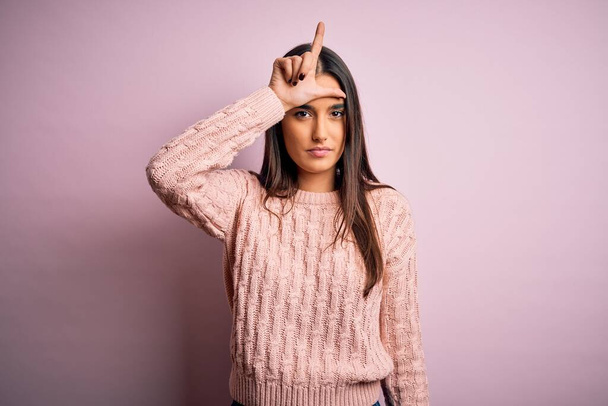 Young beautiful brunette woman wearing casual sweater over isolated pink background making fun of people with fingers on forehead doing loser gesture mocking and insulting. - Photo, Image