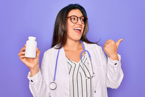 Young professional doctor woman holding pharmaceutical pills over purple background pointing and showing with thumb up to the side with happy face smiling - Photo, Image