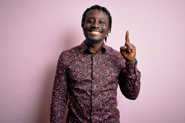 Młody, przystojny, afrykański mężczyzna ubrany w luźną koszulę, stojący nad różowym tłem i wskazujący palcem numer jeden, uśmiechnięty, pewny siebie i szczęśliwy.. - Zdjęcie, obraz