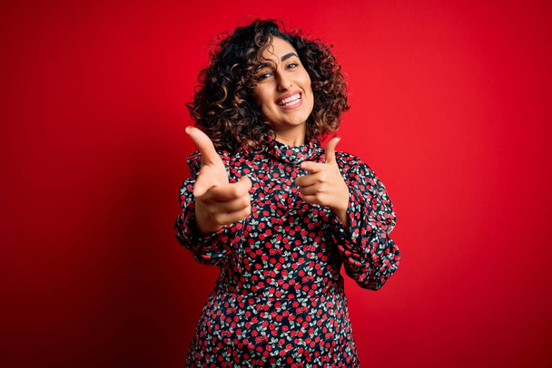 Jonge mooie krullende arabische vrouw draagt casual bloemenjurk staan over rode achtergrond wijzende vingers naar camera met gelukkig en grappig gezicht. Goede energie en vibraties. - Foto, afbeelding