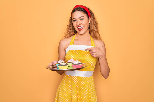 Bella pin-up donna con gli occhi azzurri che tengono i cupcake al cioccolato su sfondo giallo molto felice puntando con mano e dito
 - Foto, immagini