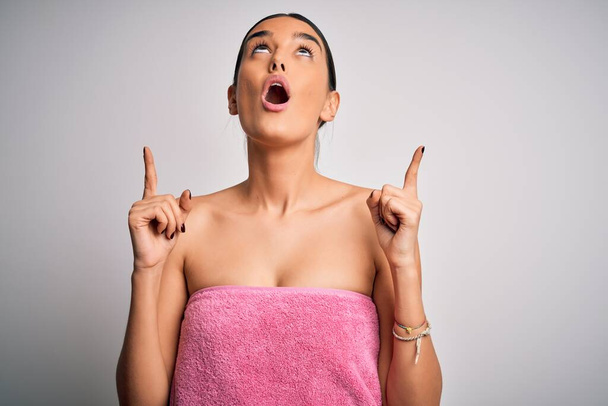 Młoda piękna brunetka kobieta nosi ręcznik po prysznicu na odizolowanym białym tle zdumiony i zaskoczony patrząc w górę i wskazując palcami i podniesionymi ramionami. - Zdjęcie, obraz