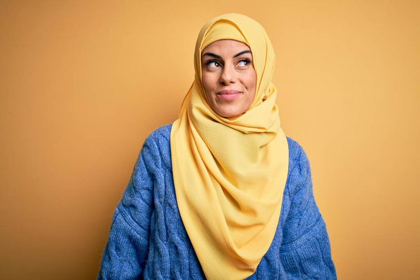 Junge schöne brünette Muslimin mit arabischem Hijab vor isoliertem gelben Hintergrund, lächelnd zur Seite schauend und denkend wegstarrend. - Foto, Bild