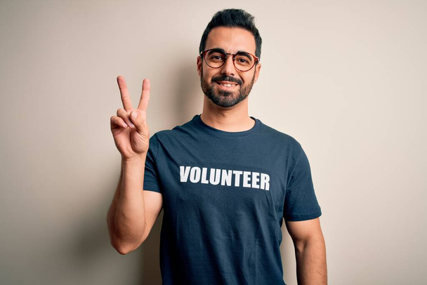 Красавчик с бородой в футболке с волонтерским посланием на белом фоне улыбается, глядя в камеру, показывая пальцы, делающие знак победы. Номер два:
. - Фото, изображение