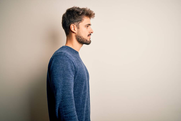 Junger gutaussehender Mann mit Bart trägt lässigen Pullover, der über weißem Hintergrund steht und zur Seite schaut, entspannte Profil-Pose mit natürlichem Gesicht mit selbstbewusstem Lächeln. - Foto, Bild