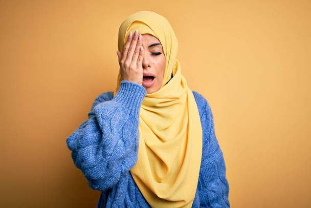 Jovem mulher muçulmana morena bonita vestindo hijab árabe sobre fundo amarelo isolado bocejo cansado cobrindo metade do rosto, olho e boca com a mão. Cara dói na dor
. - Foto, Imagem