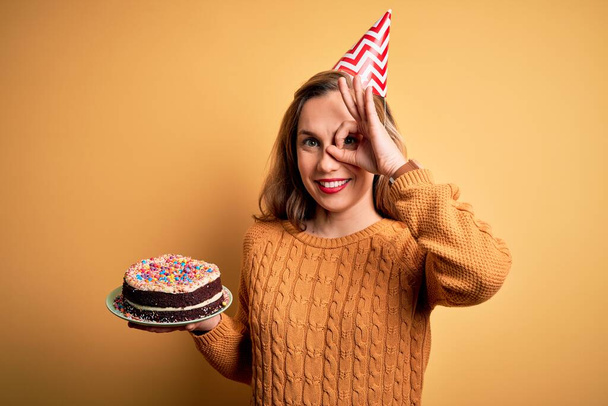 Young beautiful blonde woman holding birthday cake over isolated yellow background with happy face smiling doing ok sign with hand on eye looking through fingers - Foto, imagen
