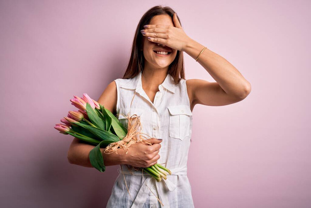 Young beautiful brunette woman holding bouquet of tulips flowers over pink background smiling and laughing with hand on face covering eyes for surprise. Blind concept. - Photo, Image